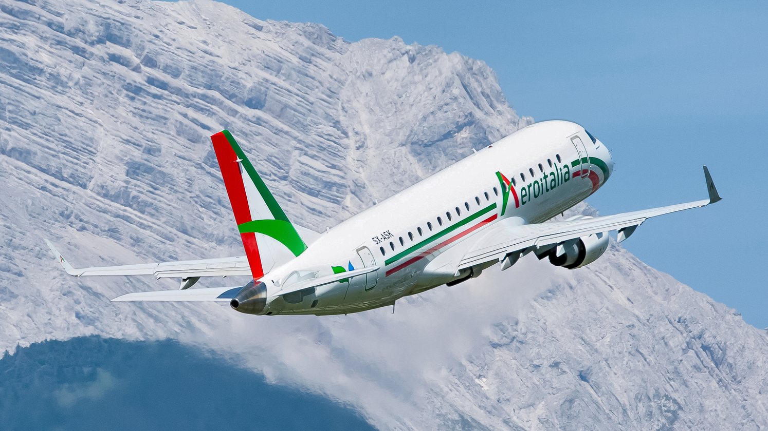 Embraer aircraft in Aeroitalia livery flying beside a snow-covered mountain